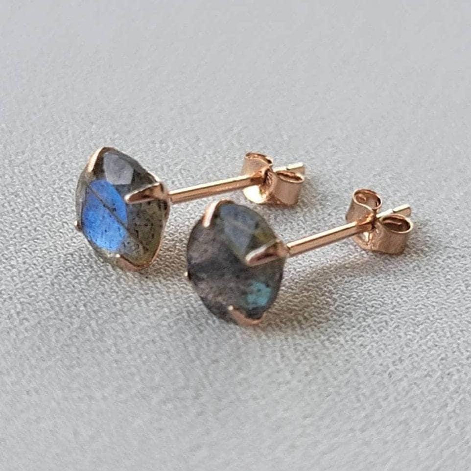 A pair of rose gold stud earrings with round, iridescent labradorite gemstones. The gemstones have a dark gray base with blue and green flashes of color.