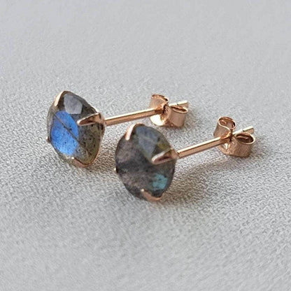 A pair of rose gold stud earrings with round, iridescent labradorite gemstones. The gemstones have a dark gray base with blue and green flashes of color.