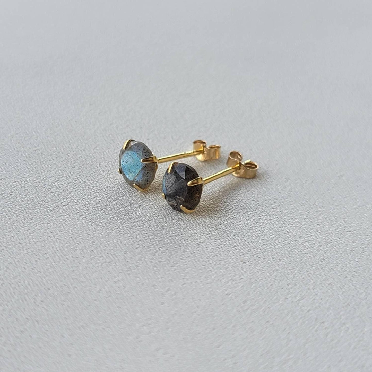A pair of gold stud earrings with round, iridescent labradorite gemstones. The gemstones have a dark gray base with blue and green flashes of color.