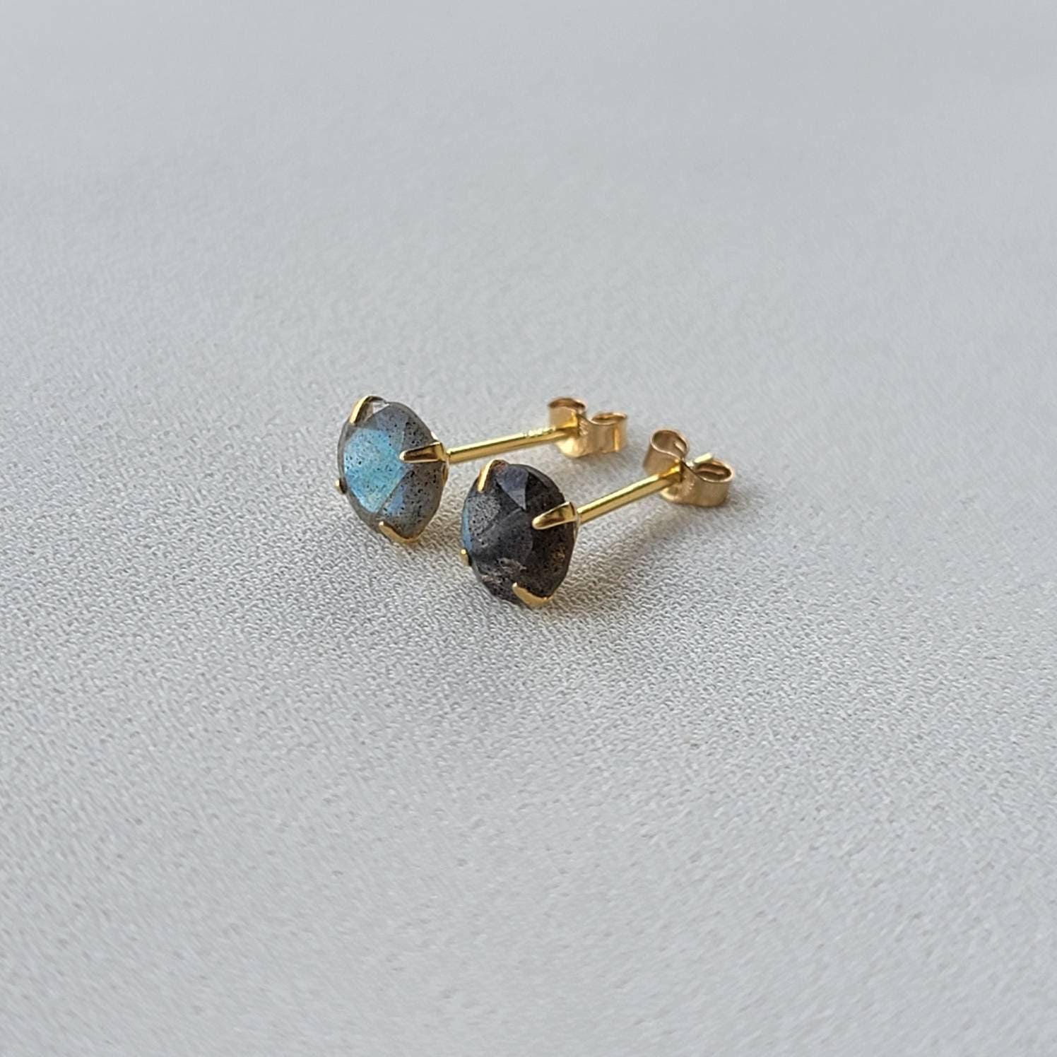A pair of gold stud earrings with round, iridescent labradorite gemstones. The gemstones have a dark gray base with blue and green flashes of color.