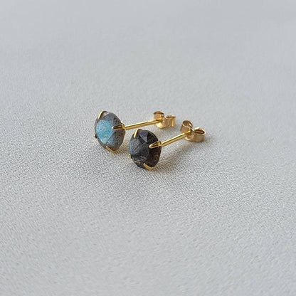 A pair of gold stud earrings with round, iridescent labradorite gemstones. The gemstones have a dark gray base with blue and green flashes of color.