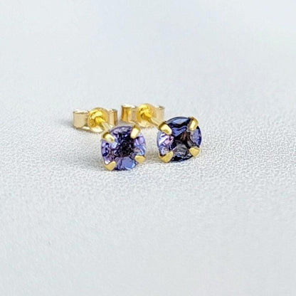 Close-up view of a pair of gold tanzanite stud earrings, each featuring a small, round, purple gemstone. The earrings have a simple, classic design, with the gemstones securely set in the gold settings. The gemstones appear to be a light to medium shade of purple. The gold is a warm, yellowish tone. The earrings are displayed against a plain, off-white background.