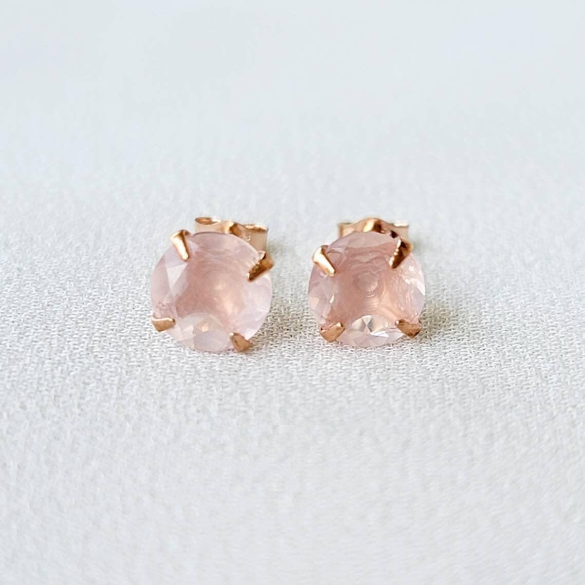 Rose gold stud earrings featuring round faceted rose quartz gemstones. Each earring showcases a light pink, translucent stone securely set in a four-prong rose gold setting. The posts and butterfly backs are also crafted from rose gold, ensuring a cohesive and elegant look. These delicate earrings are perfect for adding a touch of understated glamour to any outfit.