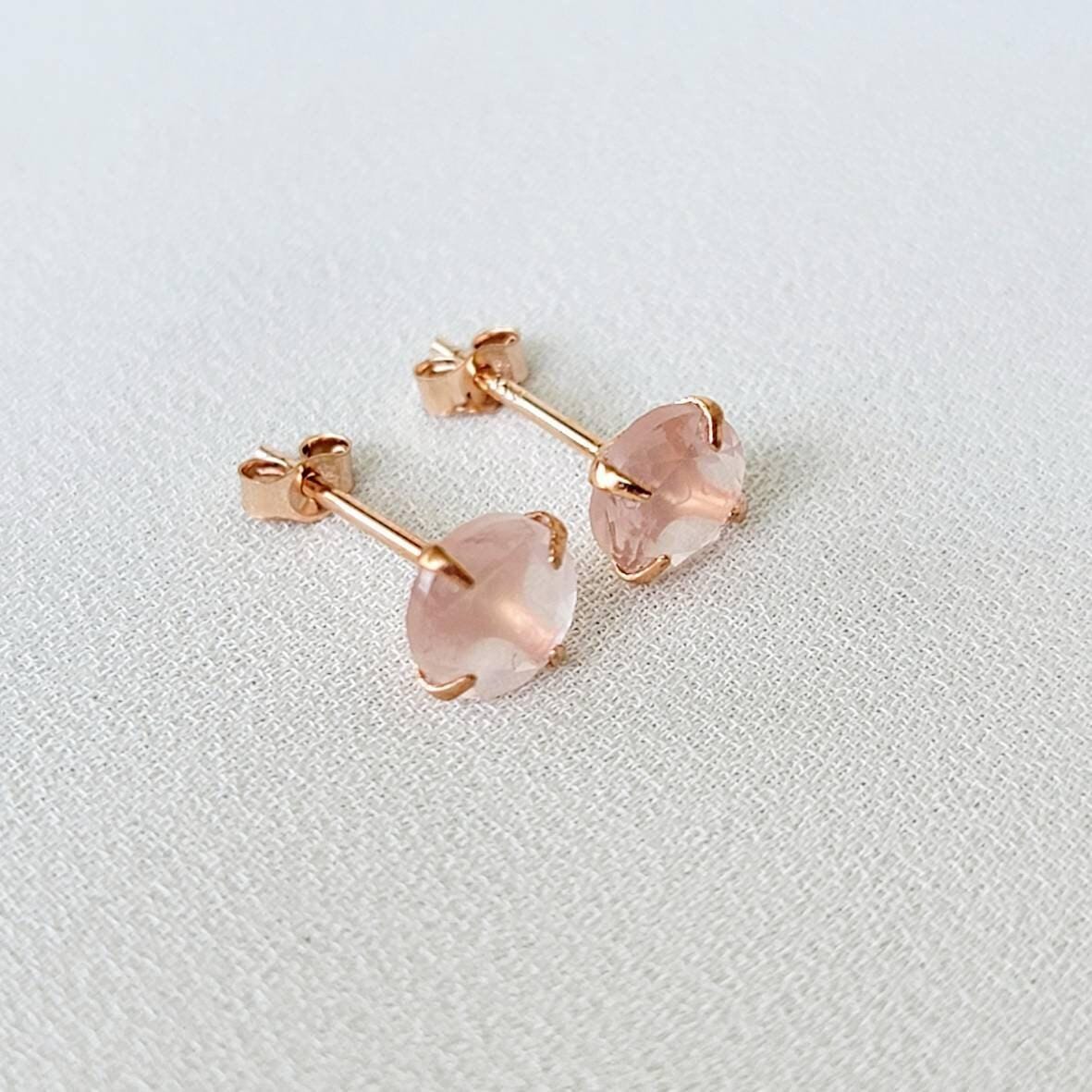 Rose gold stud earrings featuring round faceted rose quartz gemstones. Each earring showcases a light pink, translucent stone securely set in a four-prong rose gold setting. The posts and butterfly backs are also crafted from rose gold, ensuring a cohesive and elegant look. These delicate earrings are perfect for adding a touch of understated glamour to any outfit.