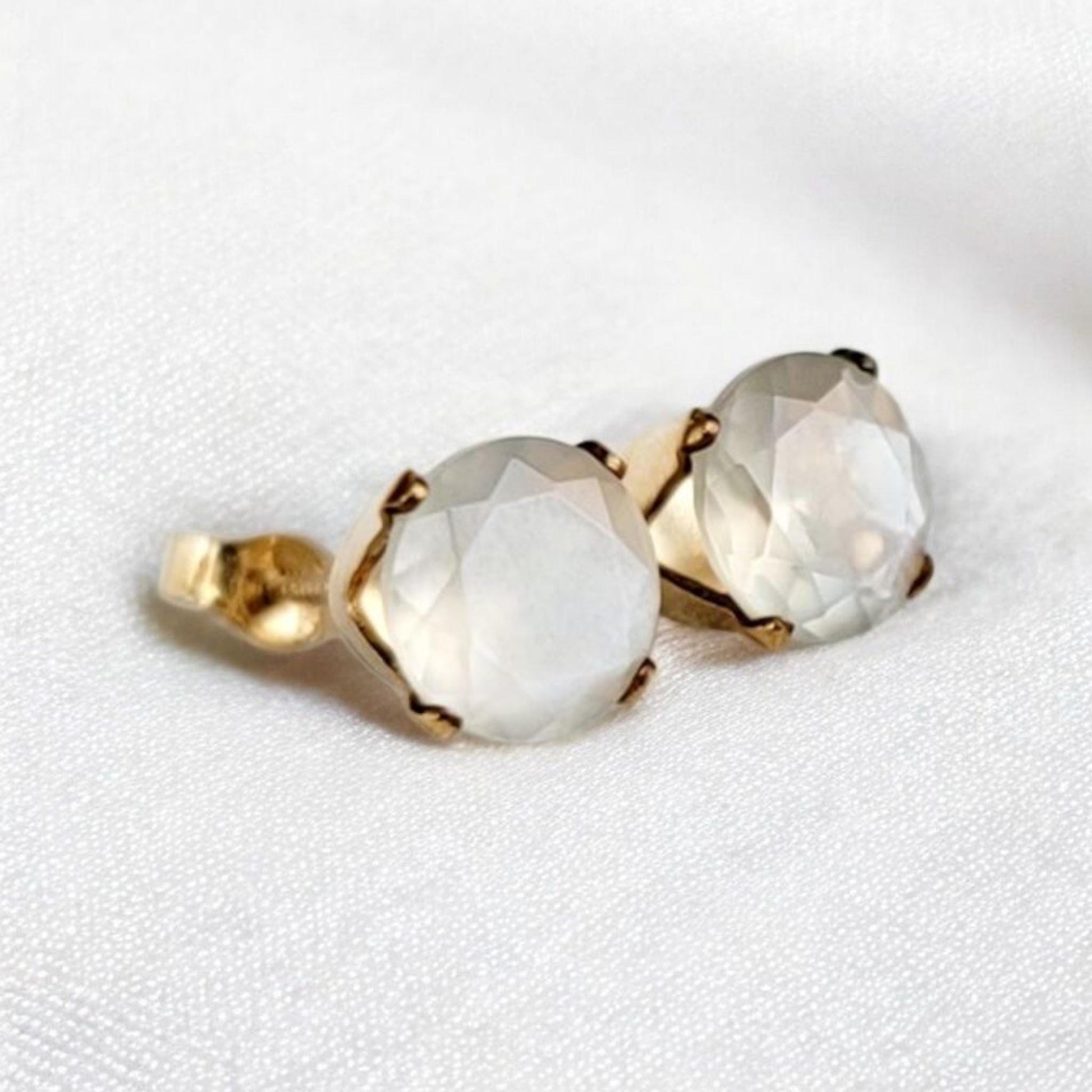 Close-up of a pair of gold stud earrings featuring round, faceted, shimmery gray moonstone gemstones. The earrings have a simple, elegant design, with the gemstones securely set in gold prongs. The gemstones have a subtle shimmer and appear to be translucent. These minimalist earrings would be a delicate addition to any jewelry collection.