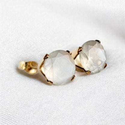 Close-up of a pair of gold stud earrings featuring round, faceted, shimmery gray moonstone gemstones. The earrings have a simple, elegant design, with the gemstones securely set in gold prongs. The gemstones have a subtle shimmer and appear to be translucent. These minimalist earrings would be a delicate addition to any jewelry collection.