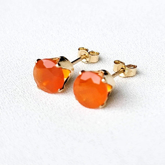 A pair of gold stud earrings with orange carnelian gemstones. The earrings have a simple design with a round gemstone set in a gold setting.