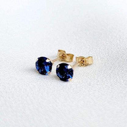 A pair of gold stud earrings with round faceted blue sapphire gemstones. The earrings are on a white surface.