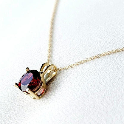 A gold chain necklace with a pendant featuring a round-shaped, dark red gemstone. The pendant is set in a gold setting with a delicate minimalist design.