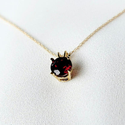A gold chain necklace with a pendant featuring a round-shaped, dark red gemstone. The pendant is set in a gold setting with a delicate minimalist design.
