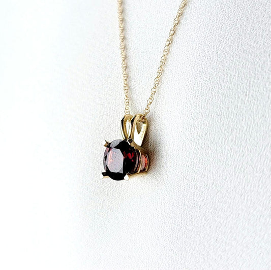 A gold chain necklace with a pendant featuring a round-shaped, dark red gemstone. The pendant is set in a gold setting with a delicate minimalist design.